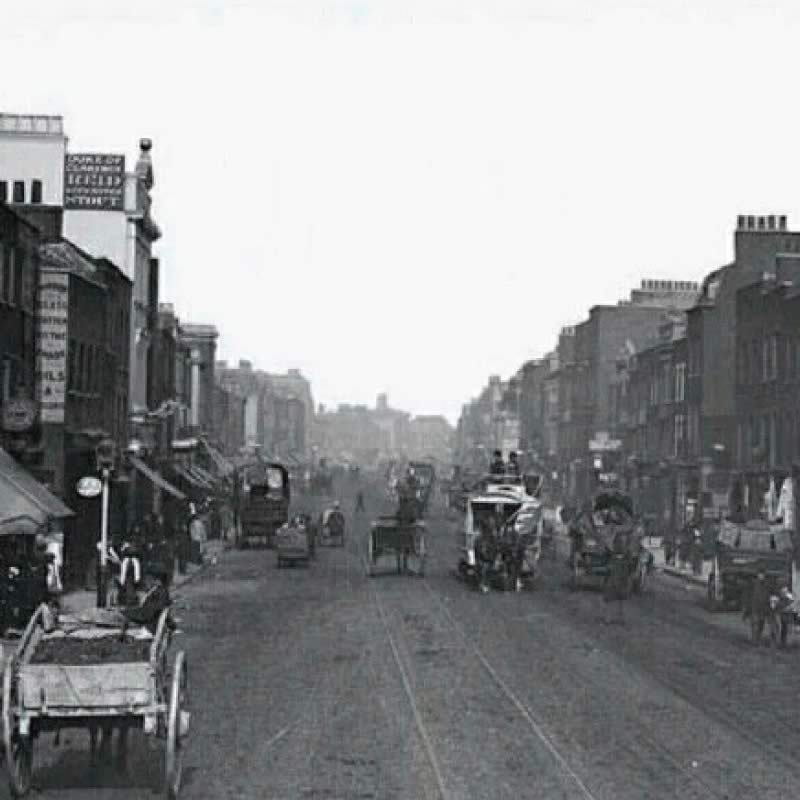 A historic black and white photo of a busy street scene with horse-drawn carriages and buildings lining both sides of the road.