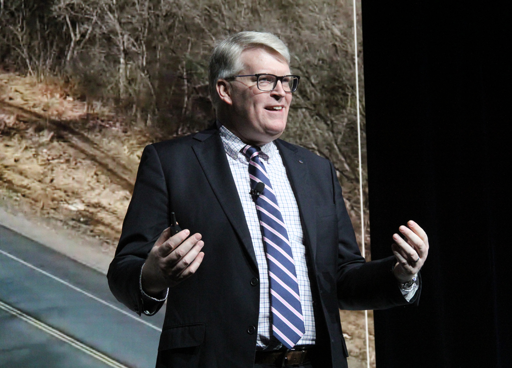 A man in a suit stands on stage, smiling and speaking with his hands raised, in front of a backdrop of a road and trees.
