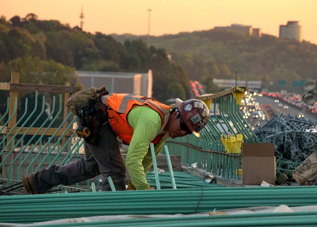 A construction worker in a safety vest and hard hat is bending over and working on metal rebar on a construction site, with a blurred cityscape in the background during sunset.
