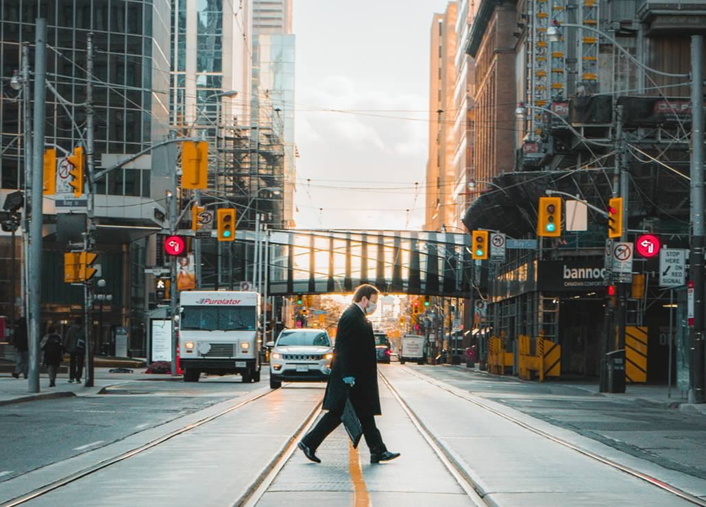A man in a black coat walking across a city street with traffic lights and buildings in the background.