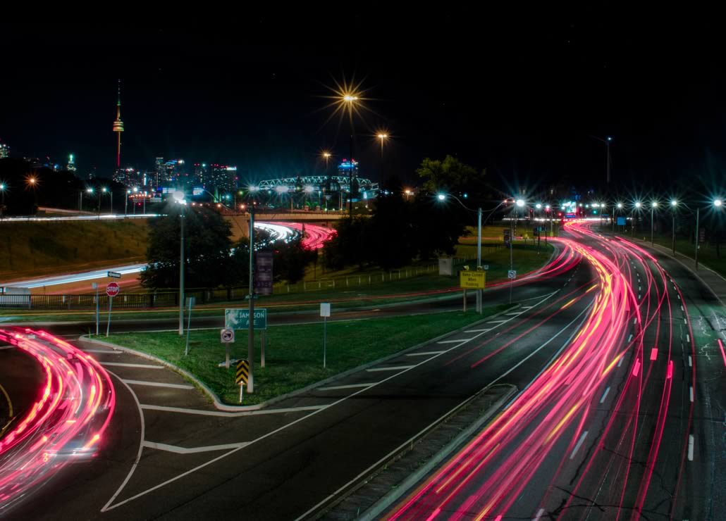 A night scene of a highway with car lights creating streaks, alongside a city skyline in the background, featuring a tower.