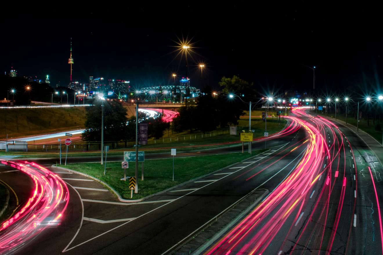 A night scene of a highway with car lights creating streaks, alongside a city skyline in the background, featuring a tower.