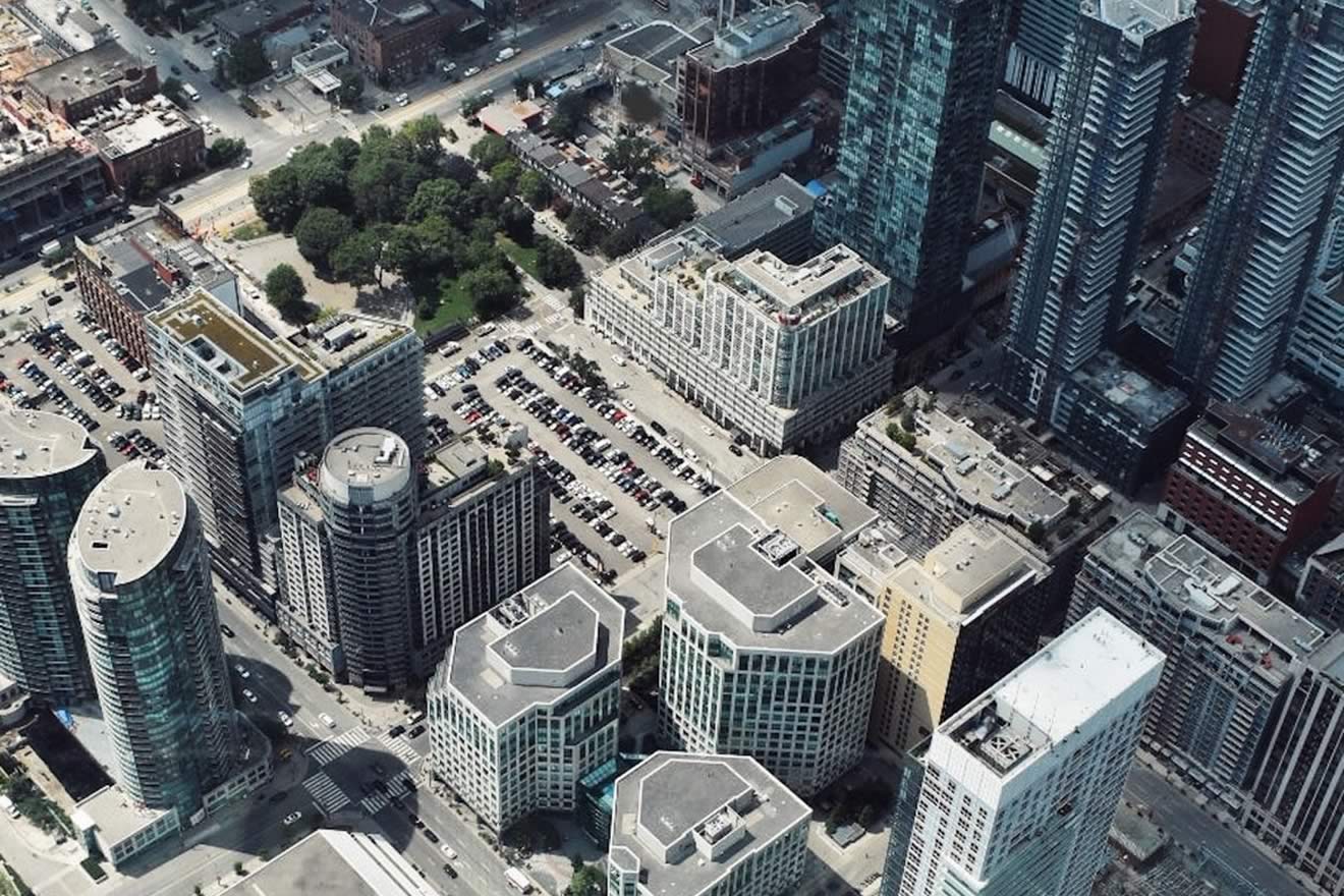 Aerial view of a cityscape featuring various buildings and a parking lot, with greenery in the distance.