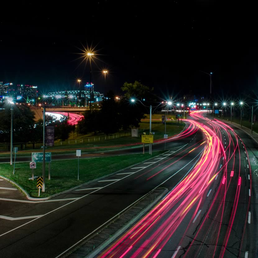 A night scene of a highway with car lights creating streaks, alongside a city skyline in the background, featuring a tower.