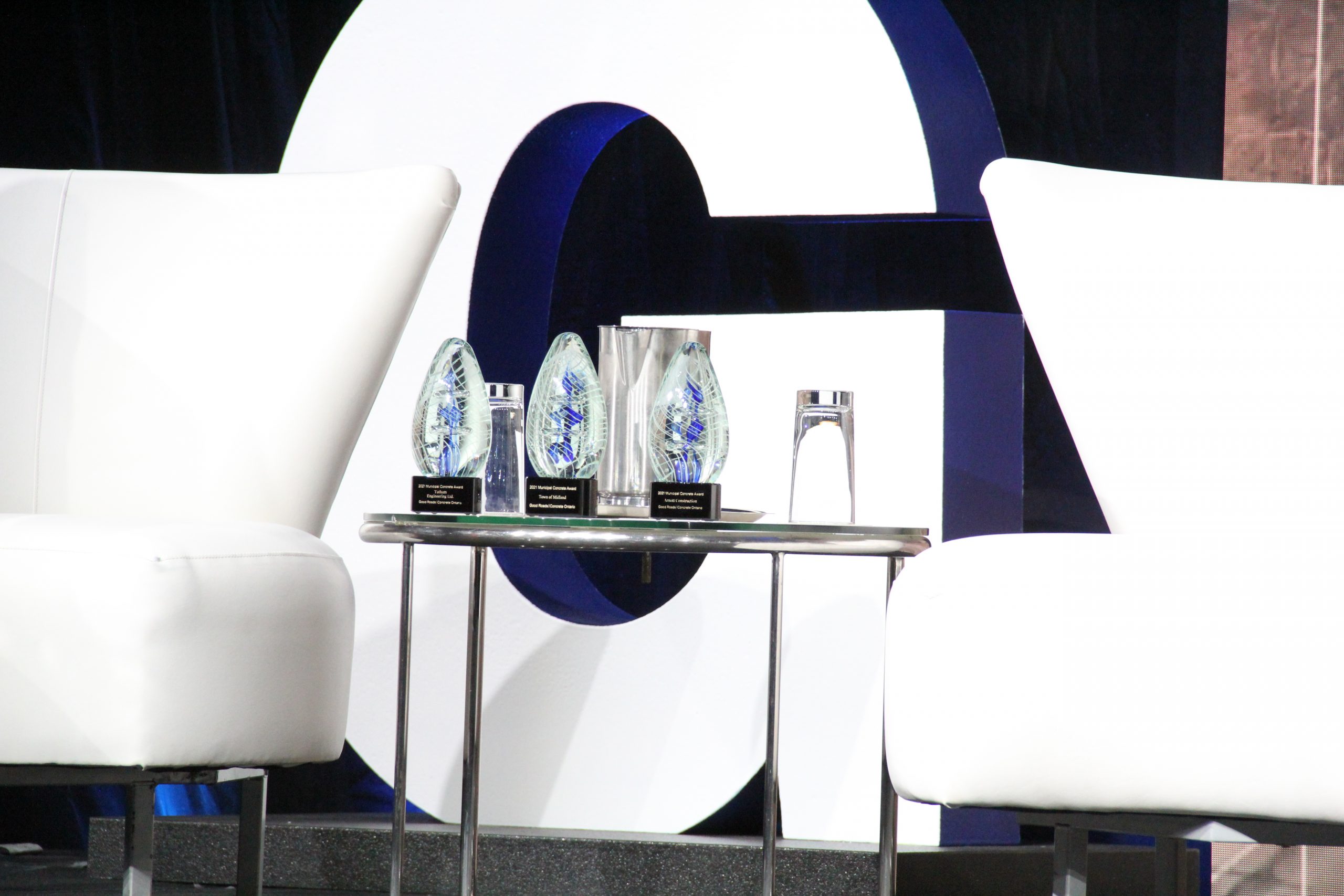 A table with several awards and a vase, beside two white chairs, with a large letter 