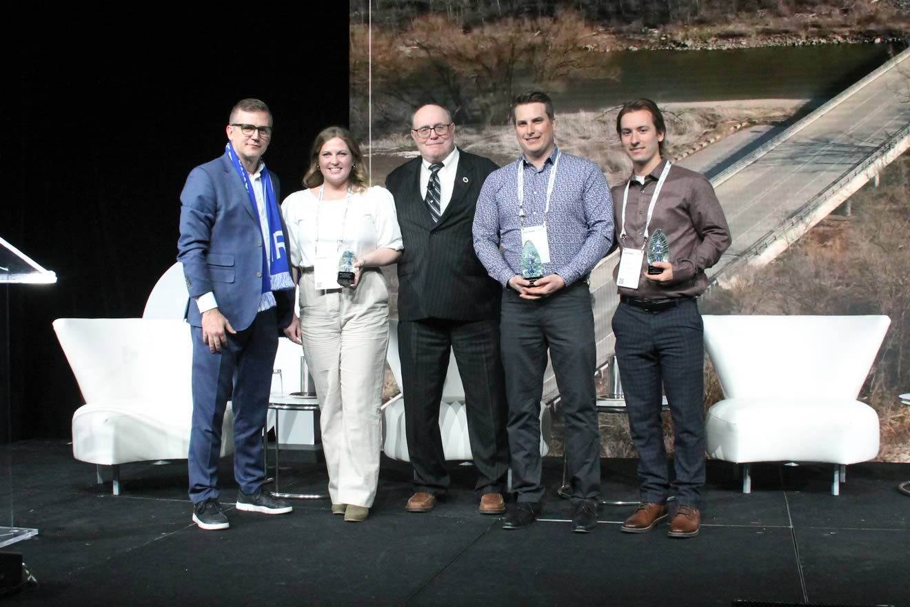 Four individuals are standing on a stage with awards in their hands, with a backdrop of a landscape featuring a bridge and a river.
