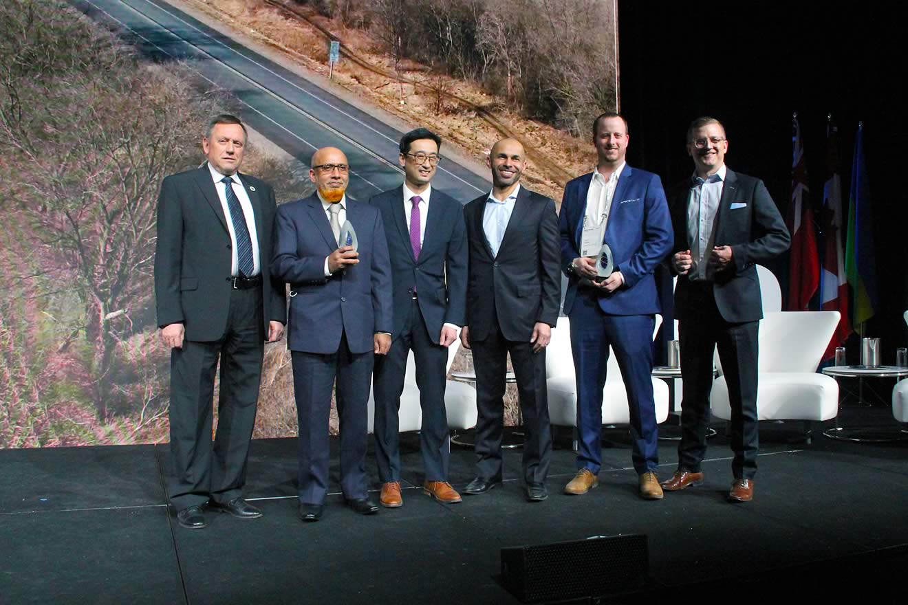 A group of six men standing together on a stage, holding awards, with a backdrop showing a road and trees.