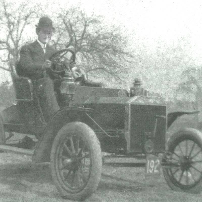 A historic black and white photo of a man in a suit and hat sitting in an early 20th-century car, with trees in the background.