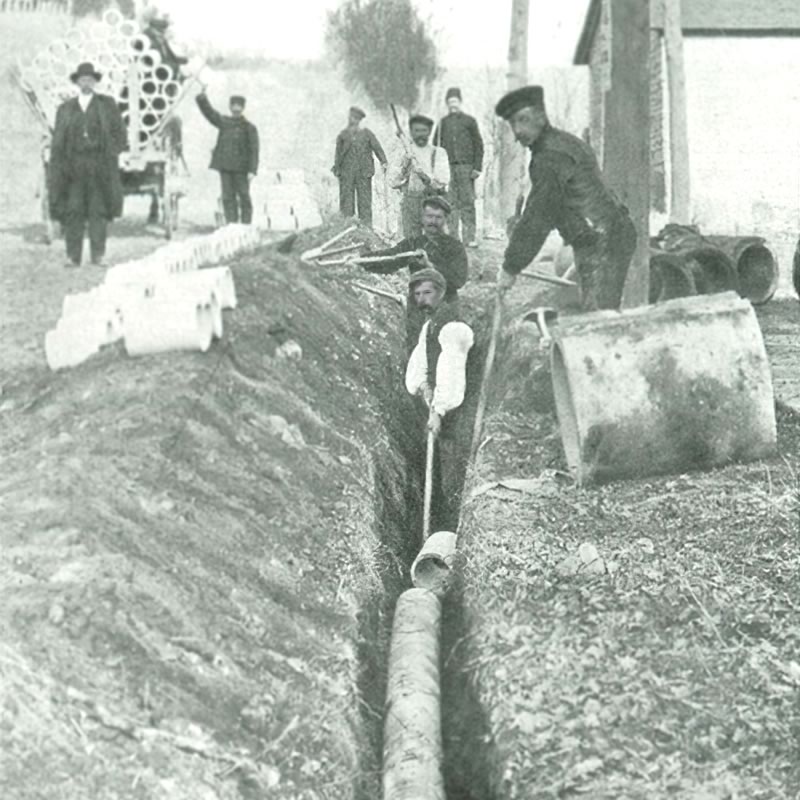 A black and white historical photograph of men working in a trench, laying pipes, with additional workers and equipment visible in the background.