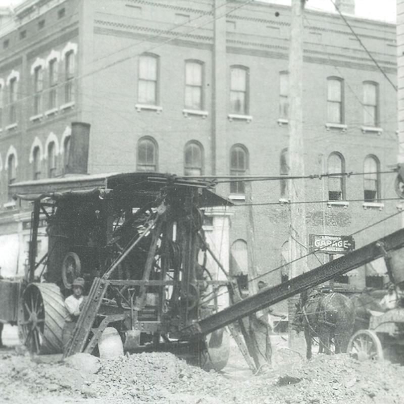 A historical black and white photo of a construction site with a large steam-powered machine and a horse-drawn wagon, set against a backdrop of brick buildings.