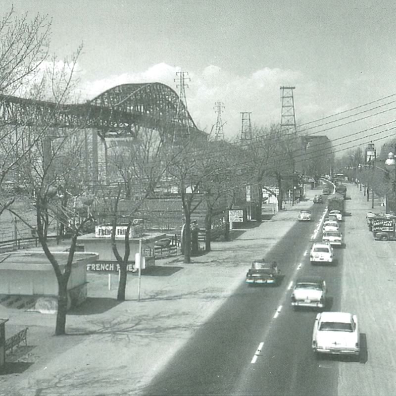 A black and white photo of a street with cars, trees, and a bridge in the background.