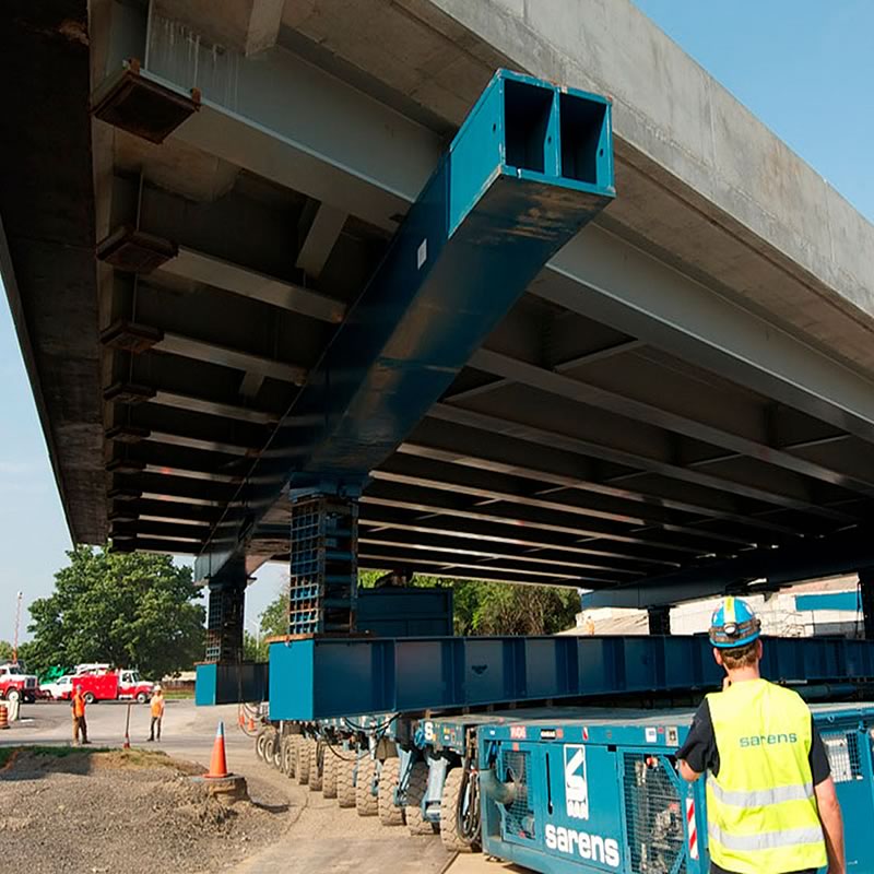 A construction worker observing a heavy blue bridge girder being positioned under a concrete bridge structure.