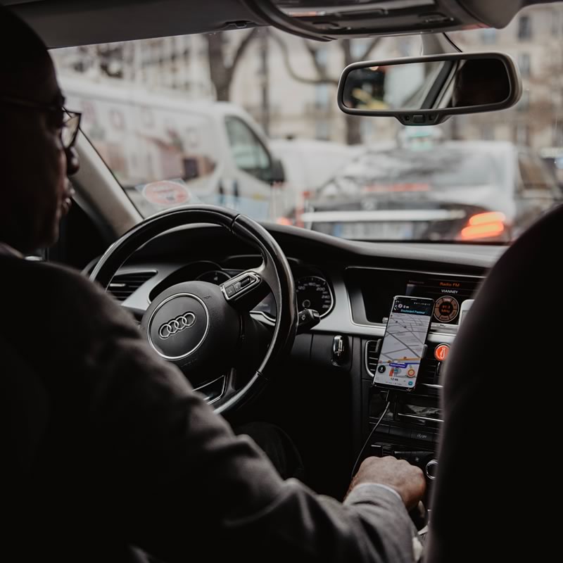A person sitting in the driver's seat of a car, looking at a mobile navigation app on a smartphone mounted on the dashboard, with traffic visible through the windshield.
