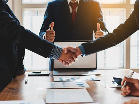 Two business people shaking hands in a meeting, with a third person giving a thumbs-up in the background. A laptop and documents are on the table.