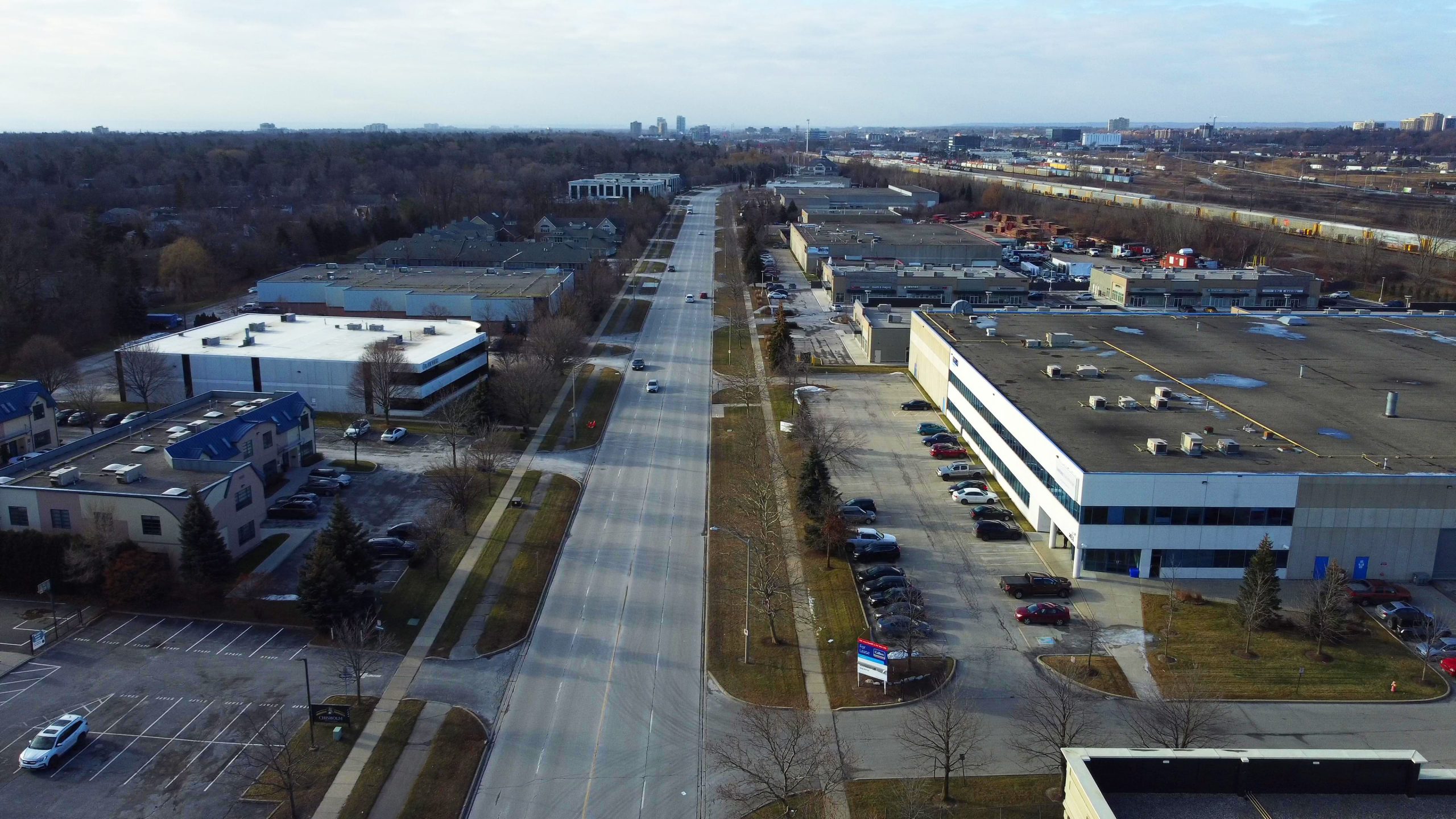 An aerial view of a wide road flanked by commercial buildings and parking lots, with a distant city skyline visible on the horizon.