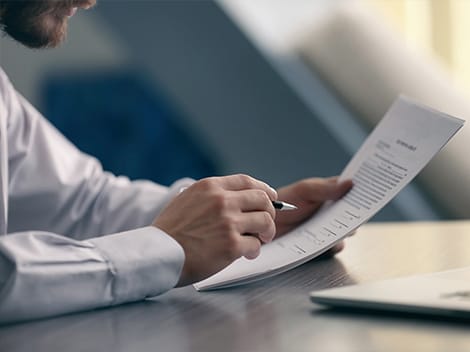 A person holding a pen and a paper while reviewing a document, with a laptop visible on the table.