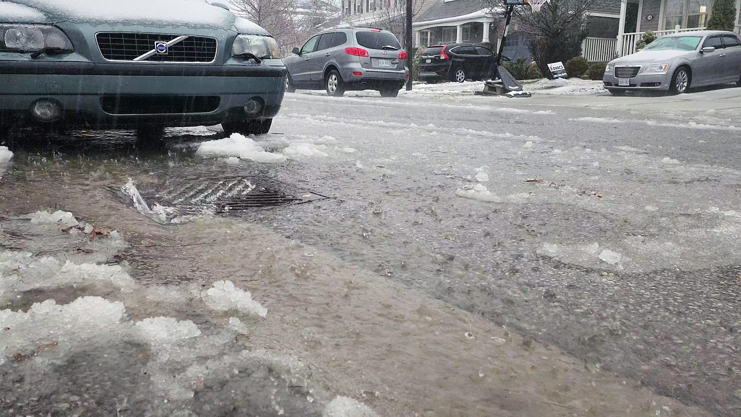 A street scene with a gray car parked on a snowy, slushy road with water pooling and flowing towards a drain.