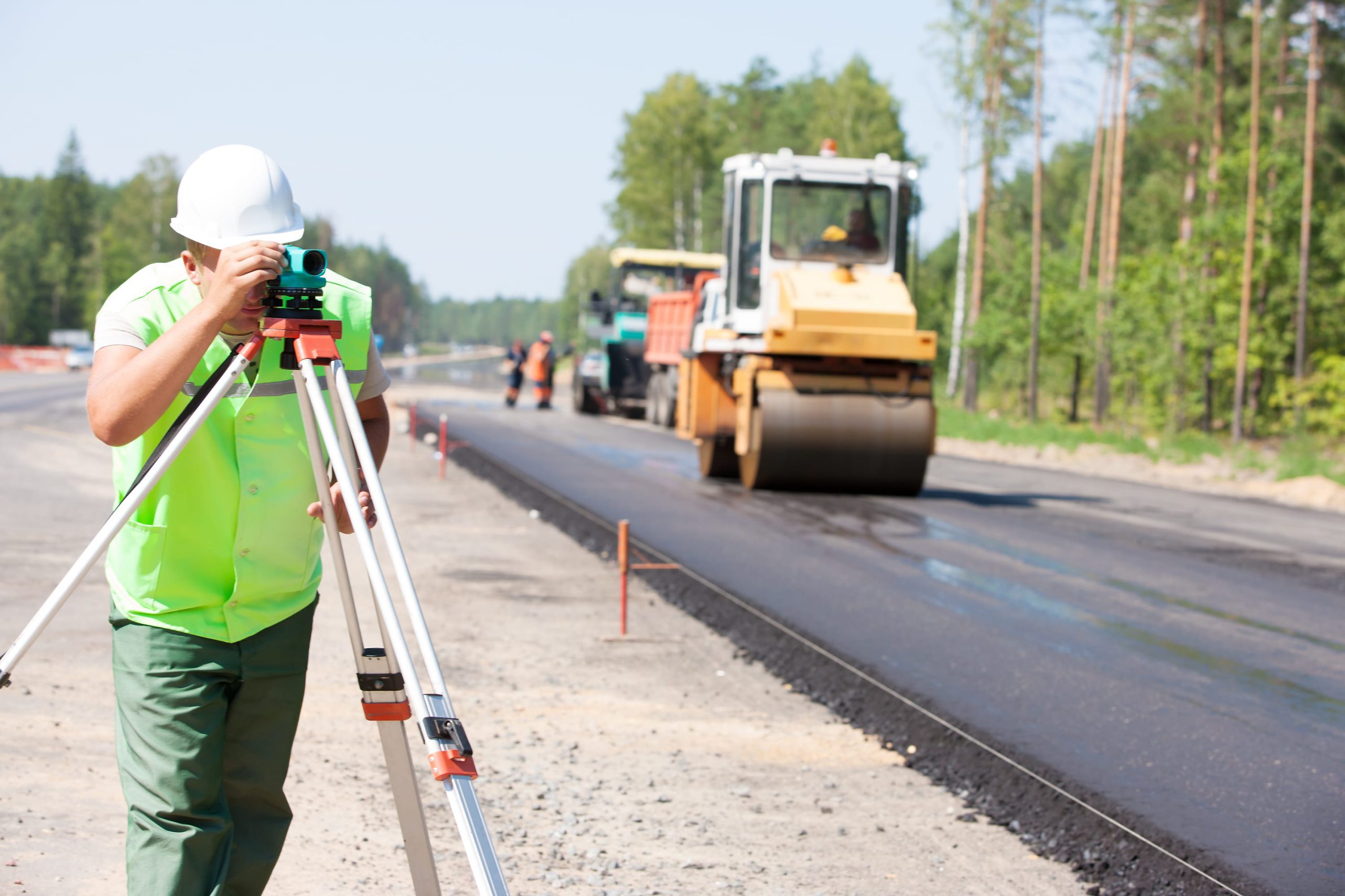 A construction worker in a green vest and white helmet uses a surveying instrument on a road construction site, with machinery and workers in the background.