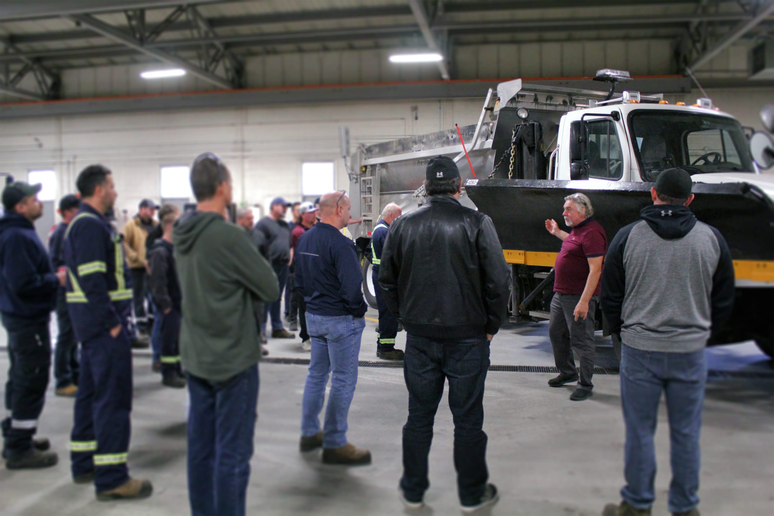 A group of people are gathered around a truck in an industrial setting, while a person gestures and speaks to them.