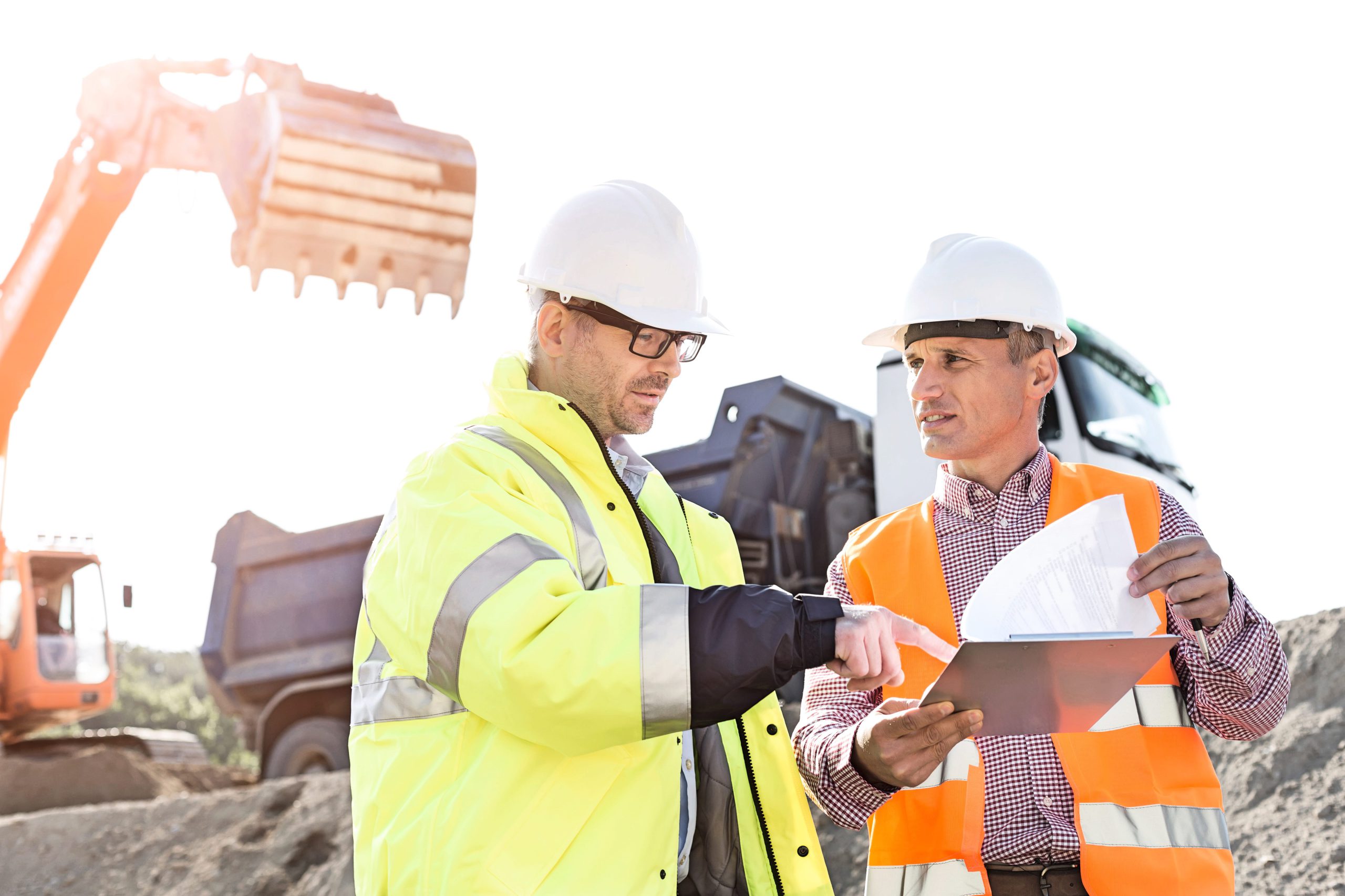 Two construction workers discussing plans on a construction site, wearing safety helmets and high-visibility jackets, with heavy machinery in the background.