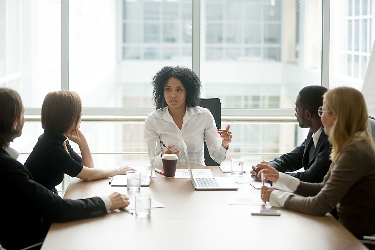 A group of professionals sitting around a conference table, attentively listening to a woman presenting with a tablet in her hand.