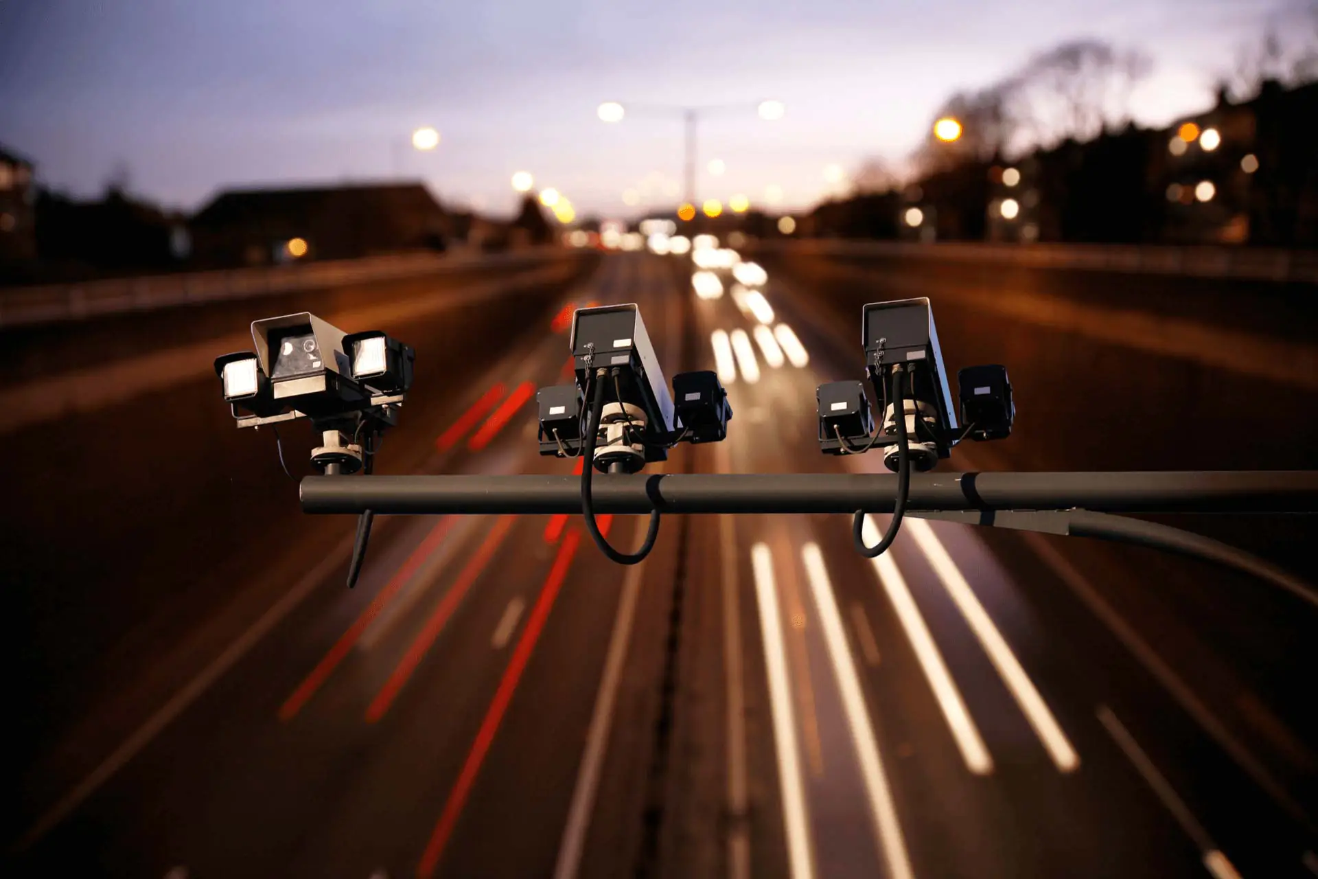 Traffic cameras mounted on a pole overlooking a highway at dusk, with light trails from vehicles moving below.