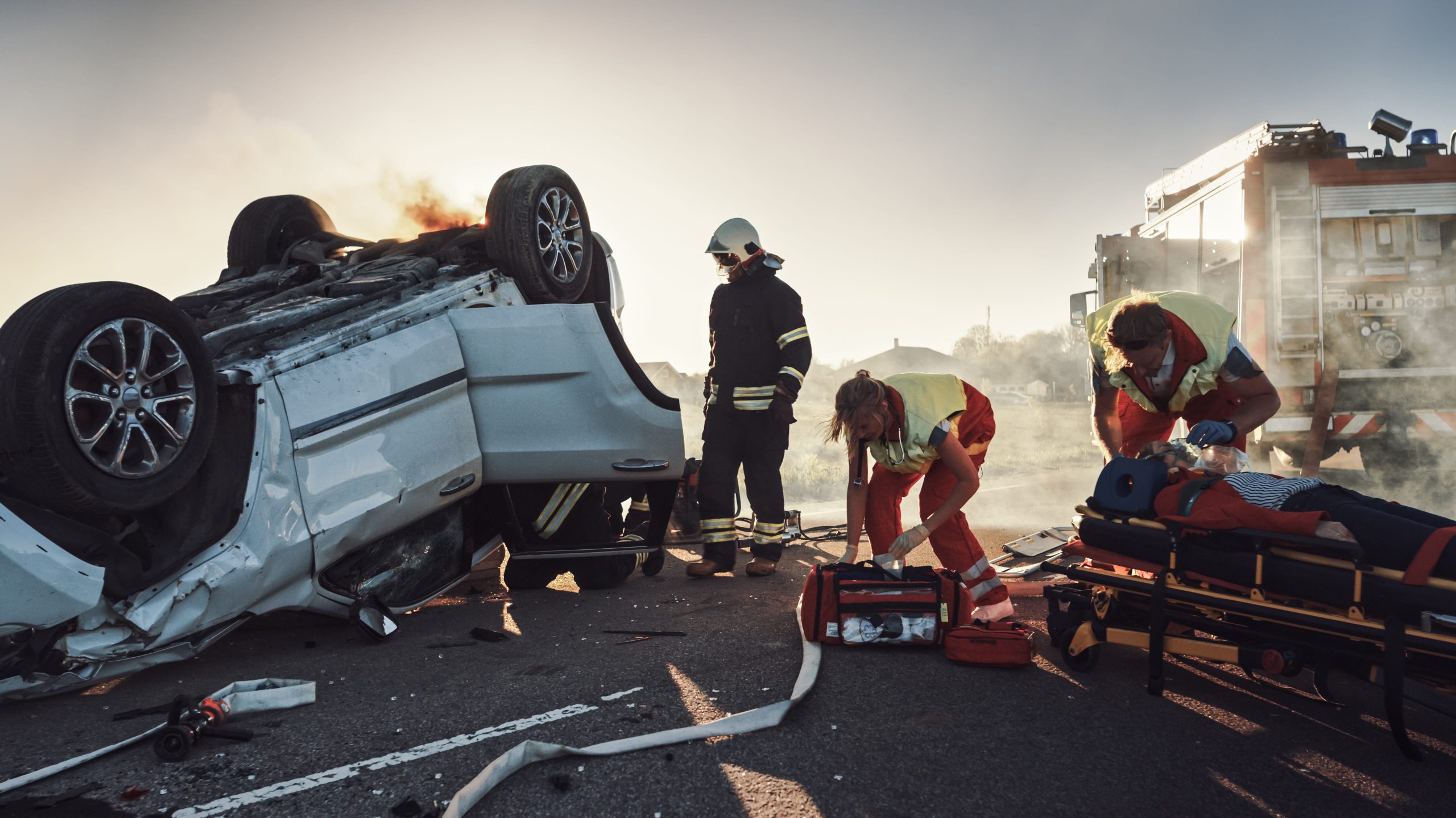 A car accident scene with an overturned vehicle, emergency responders attending to the situation, and smoke in the background.