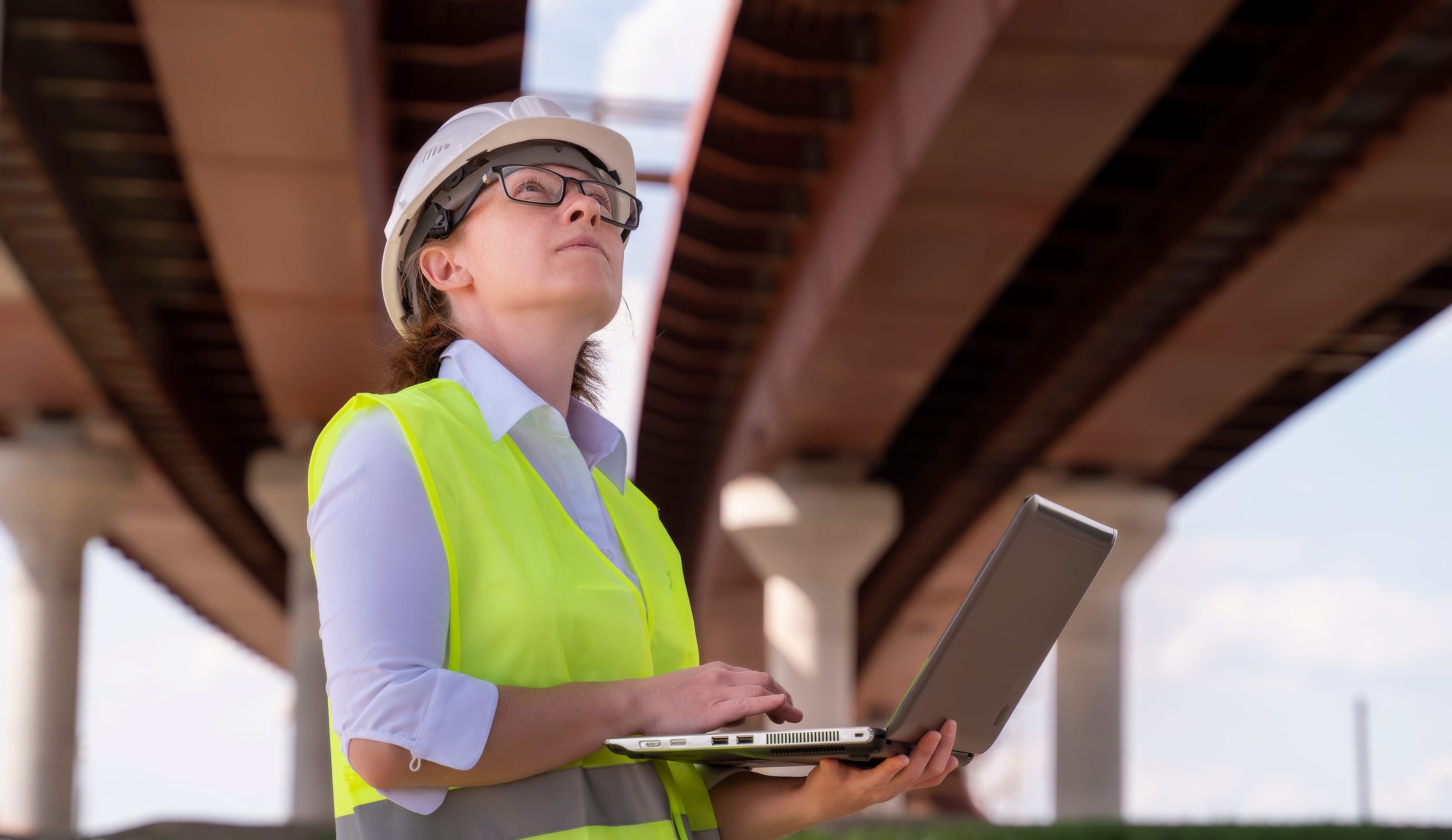 A woman in a hard hat and safety vest is working on a laptop under a bridge.
