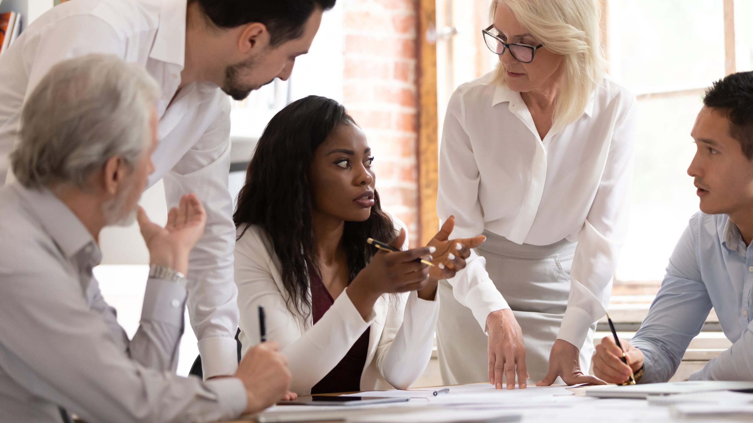 A diverse group of five colleagues engage in a focused discussion around a table, with papers and pens, conveying collaboration and teamwork in a professional setting.