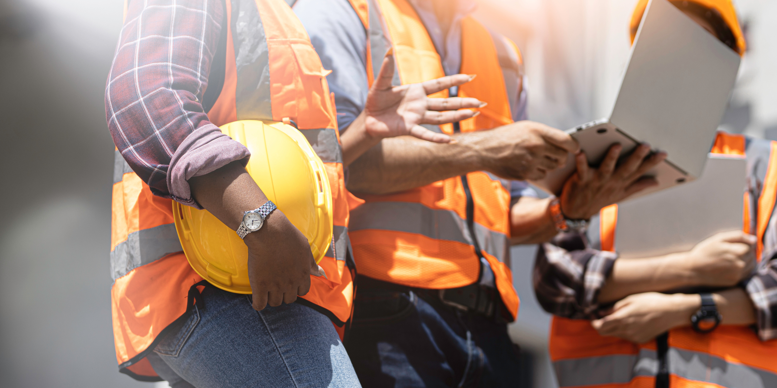 A group of construction workers in safety vests and hard hats discussing plans, with one holding a tablet and another holding a yellow hard hat.