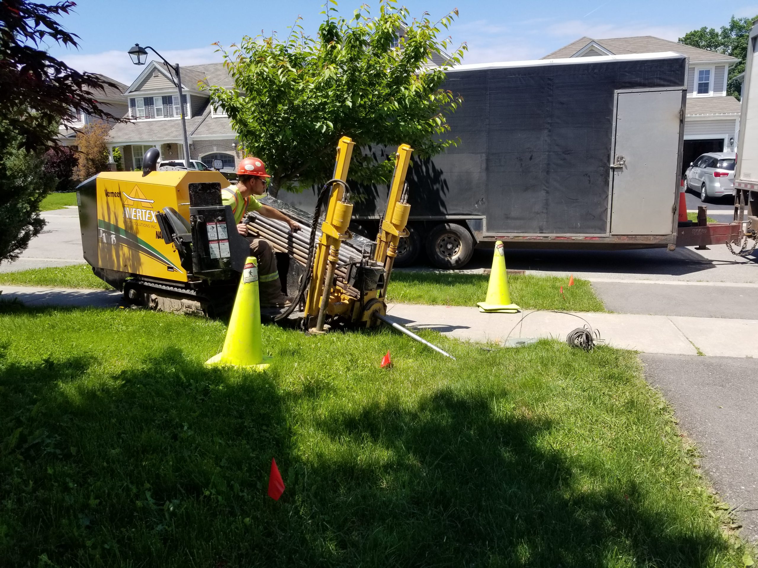 A construction worker operates a trenching machine on a grassy area near a street, with traffic cones and a trailer visible in the background.