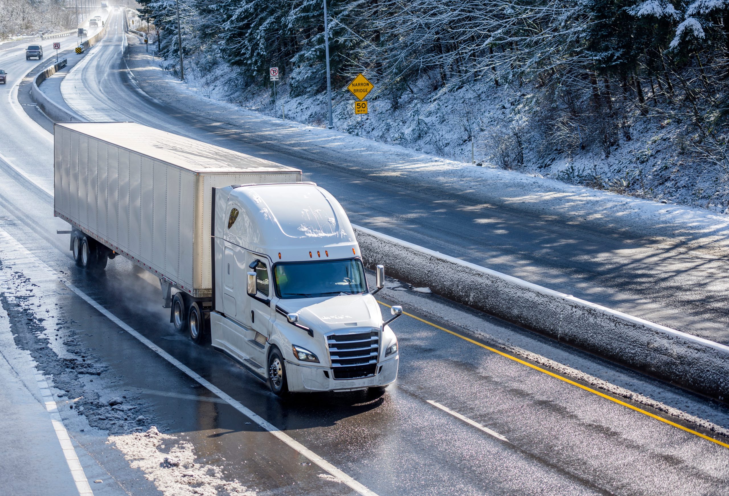 A white semi-truck with a trailer driving on a snow-covered highway surrounded by trees.