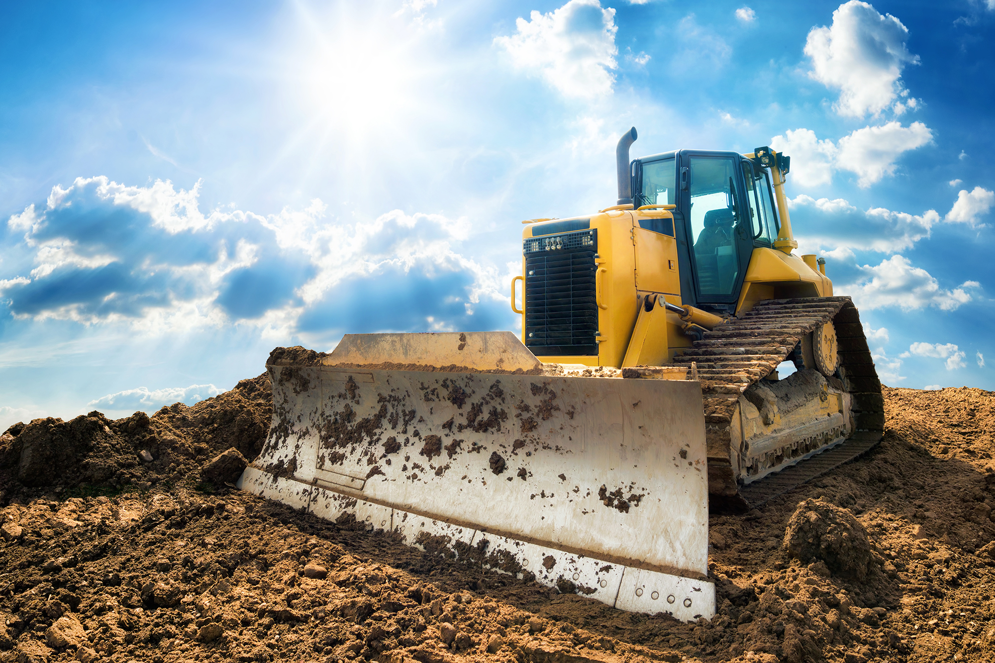 A yellow bulldozer on a construction site under a bright blue sky with fluffy clouds.