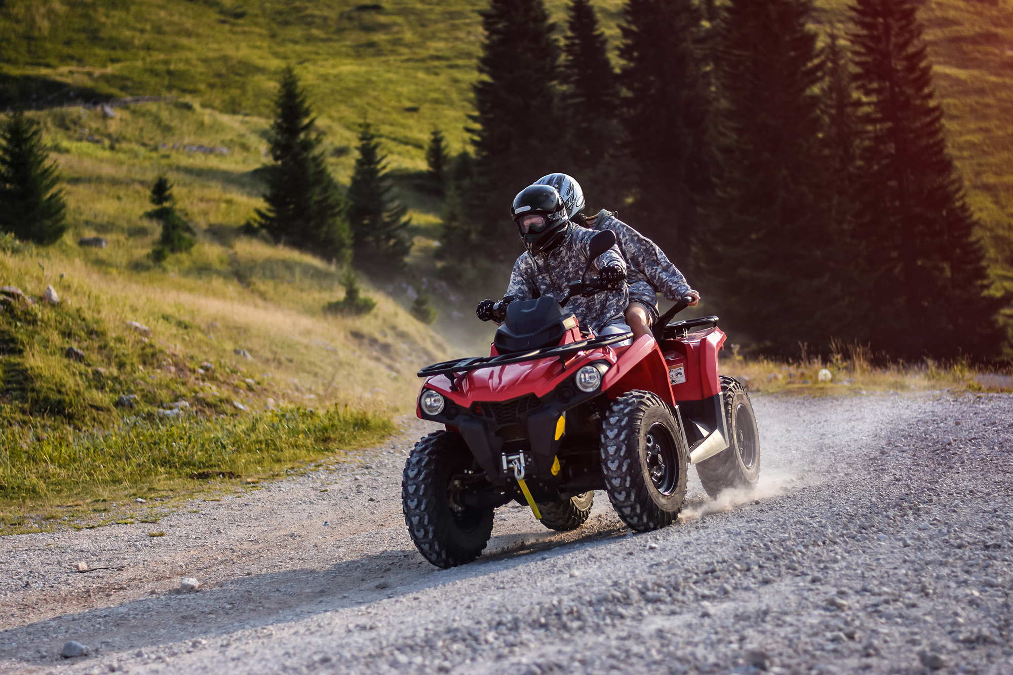 Two people riding an ATV on a gravel path surrounded by green hills and trees.