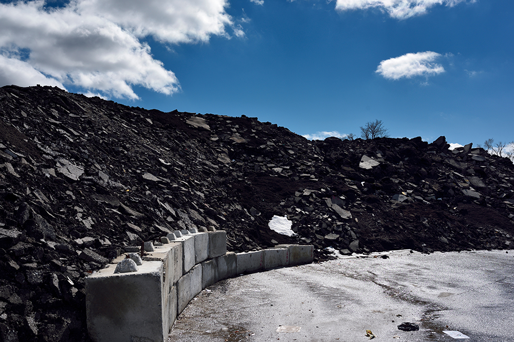A large pile of reclaimed asphalt against a blue sky with scattered clouds, beside a curved concrete barrier on a paved surface.