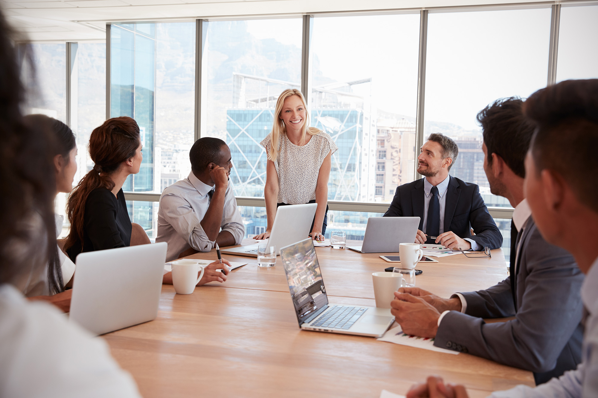 A group of professionals sitting around a conference table with laptops and notepads, engaged in a business meeting. A woman stands and speaks to the group while others listen attentively.