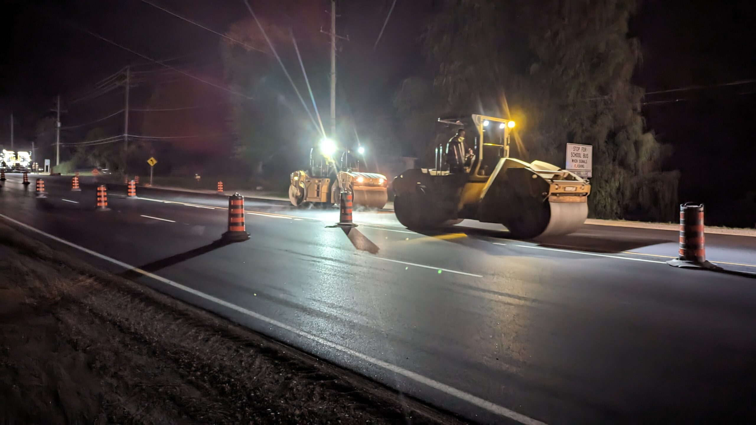 A construction scene at night with heavy machinery working on a road, surrounded by traffic cones and lights.