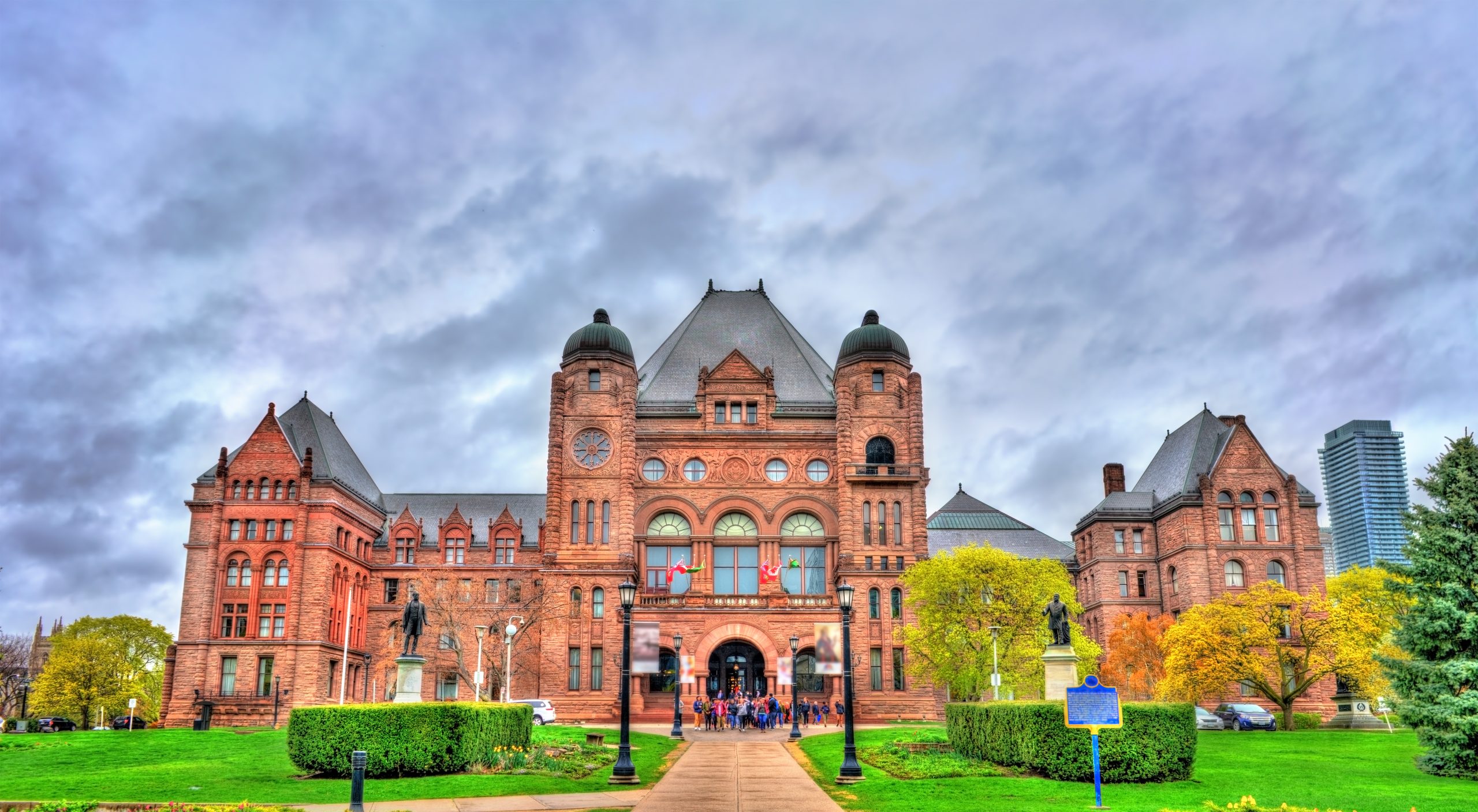 A large historic building with a red brick facade, surrounded by greenery and trees, under a cloudy sky.