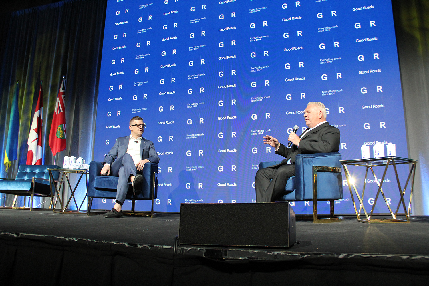 Two men seated on stage in front of a blue backdrop with logos, engaged in a discussion.