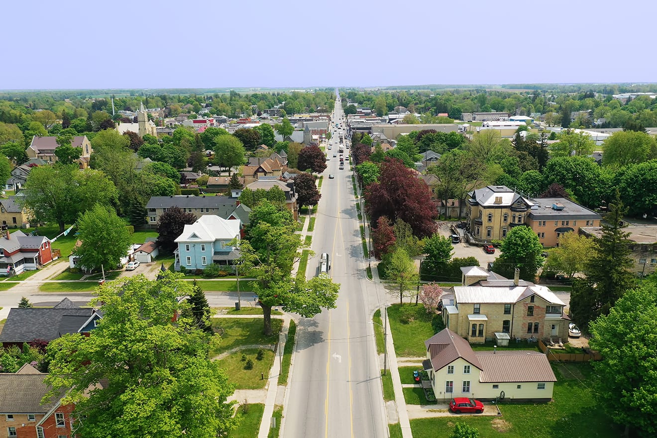 Aerial view of a suburban street lined with houses and trees, showing a long road stretching into the distance with some vehicles visible.