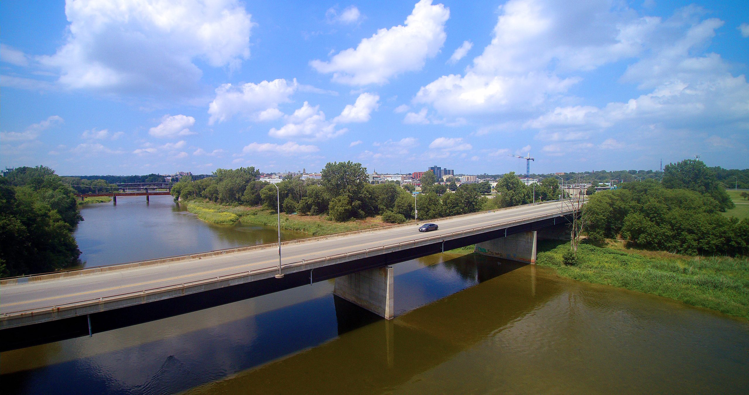 Aerial view of a highway bridge crossing over a river, with trees and buildings in the background under a partly cloudy sky.