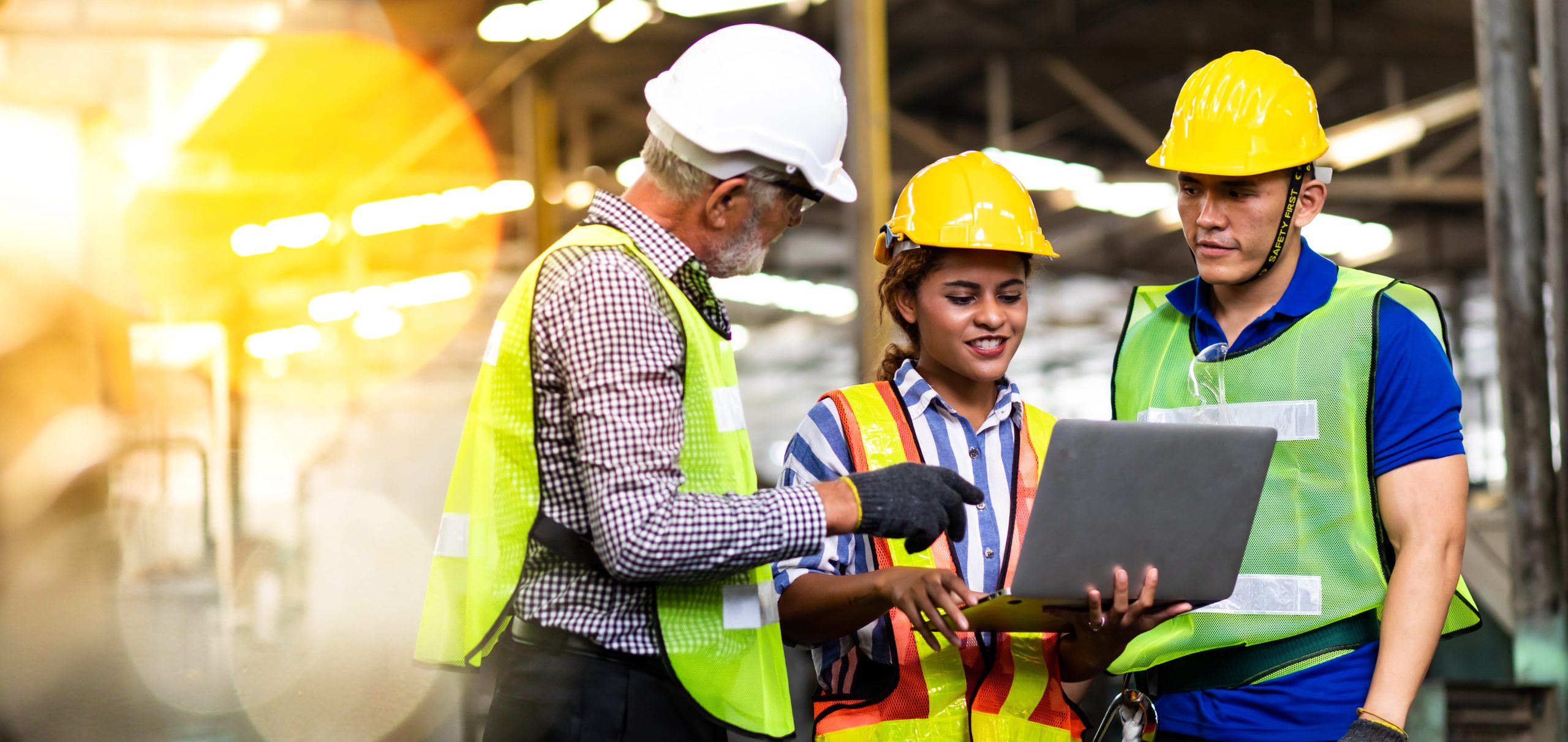 A group of three construction workers wearing hard hats and safety vests, discussing over a laptop in an industrial setting.