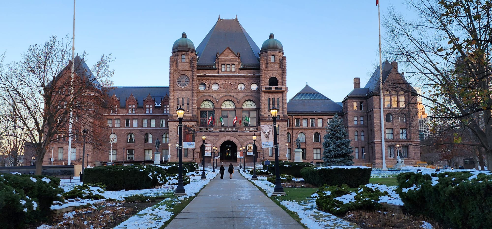 A historic building with a grand architectural design, surrounded by a pathway and landscaped grounds, showing signs of winter with some snow present.