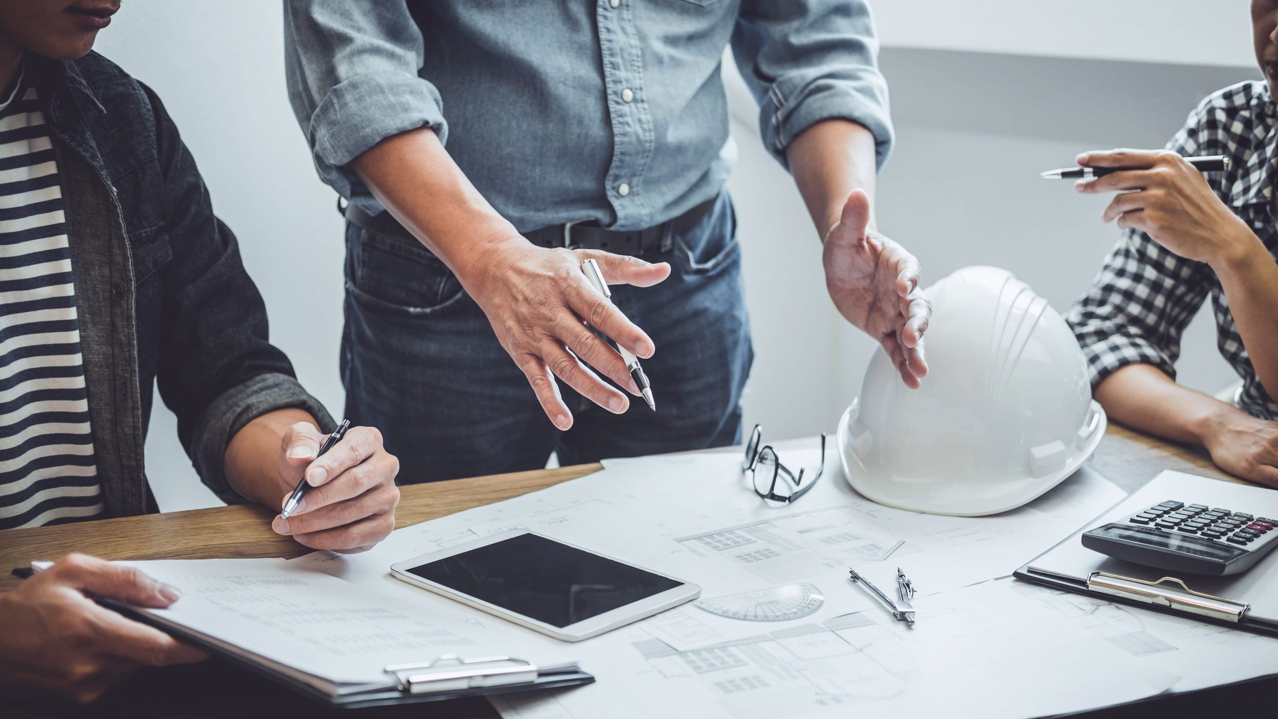 A group of people collaborating at a table, discussing plans, with a construction helmet and various stationery items.