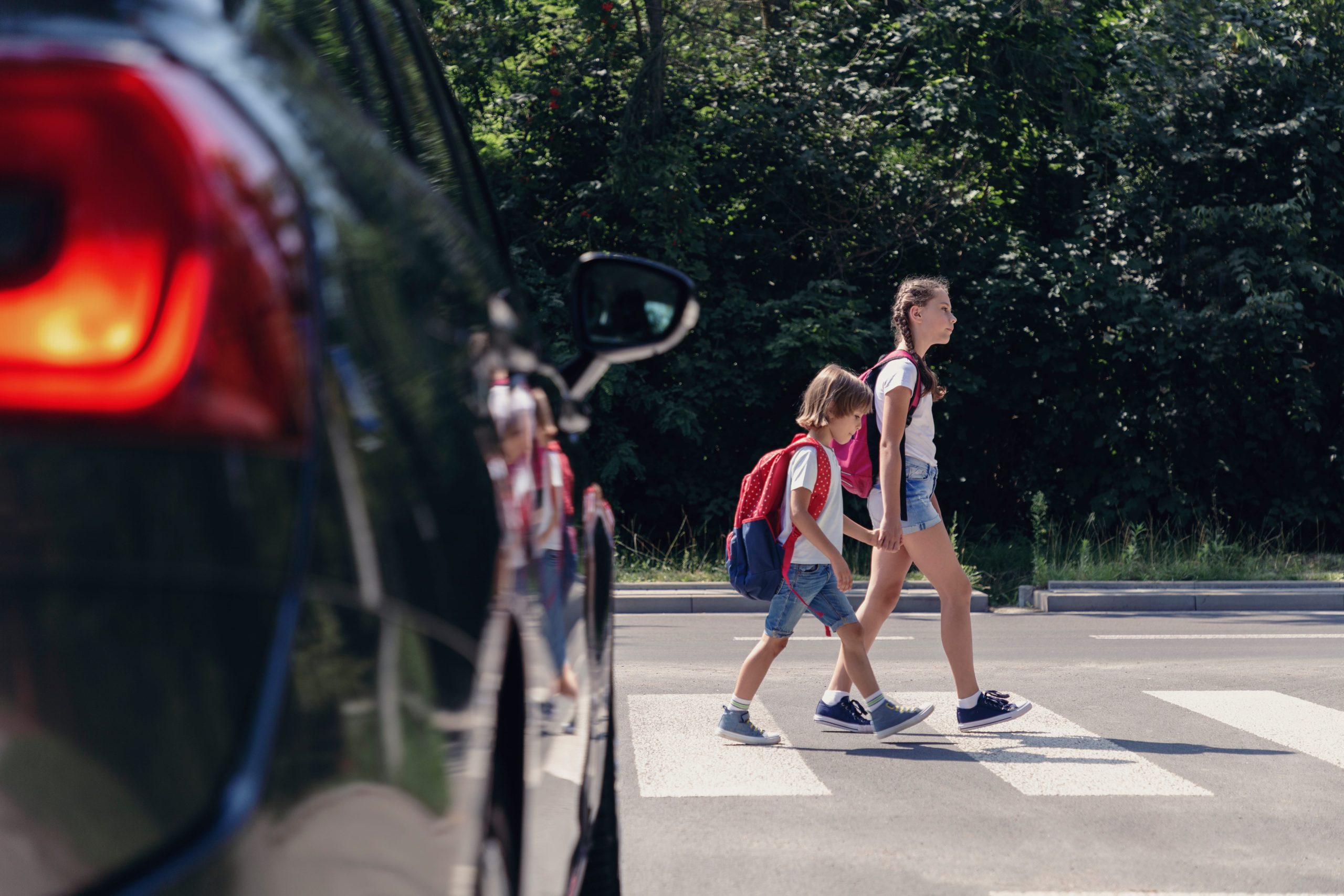 Two children with backpacks walk across a crosswalk while a car waits nearby; trees and greenery are visible in the background.