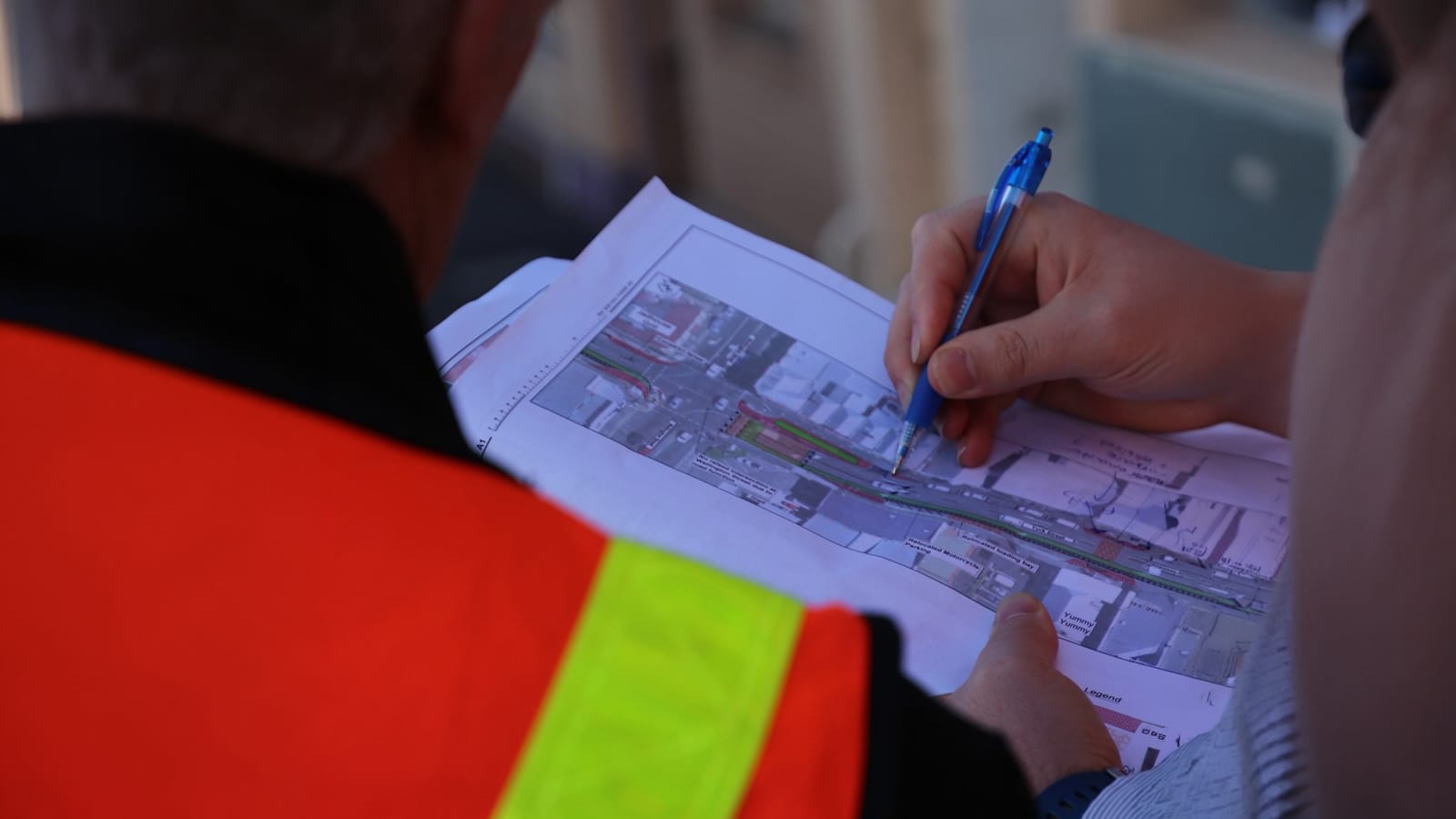 Two individuals reviewing a detailed map, one marking with a blue pen, wearing safety vests in a construction setting.