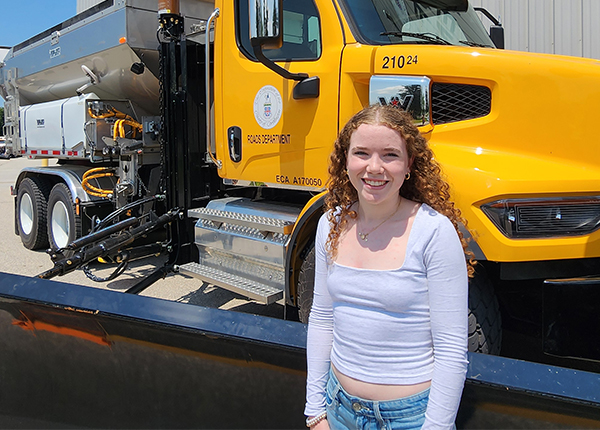 A young woman stands in front of a yellow truck with a plow attachment.