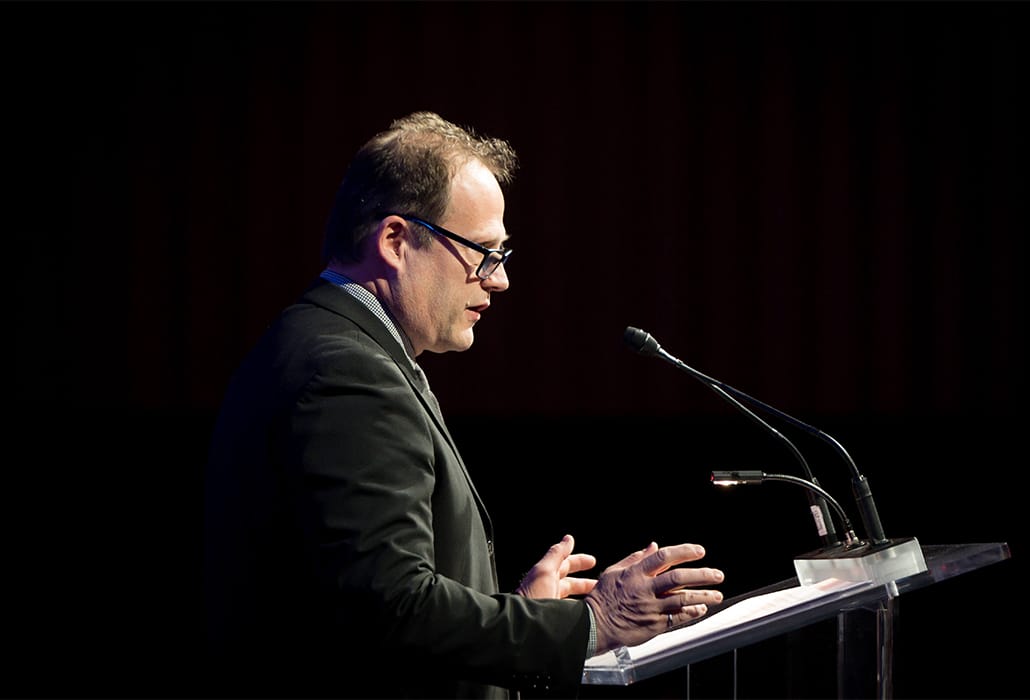 A man in a suit speaking at a podium with a microphone.