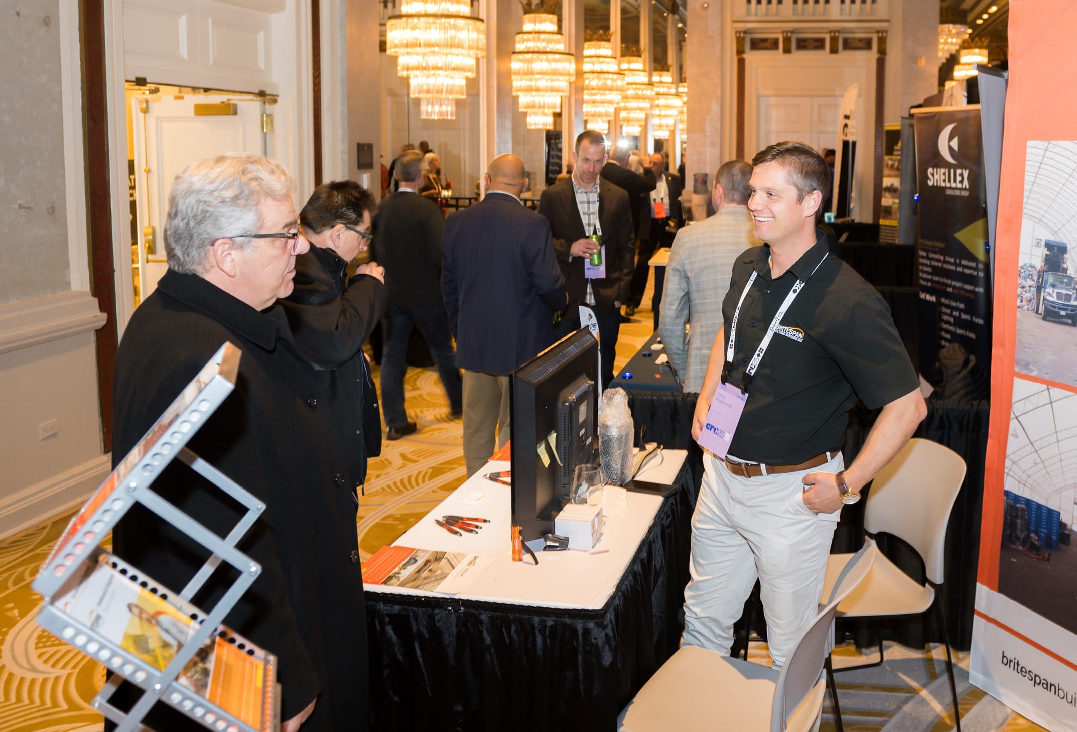 Two men are engaged in conversation at a conference or trade show, with displays and attendees in the background.