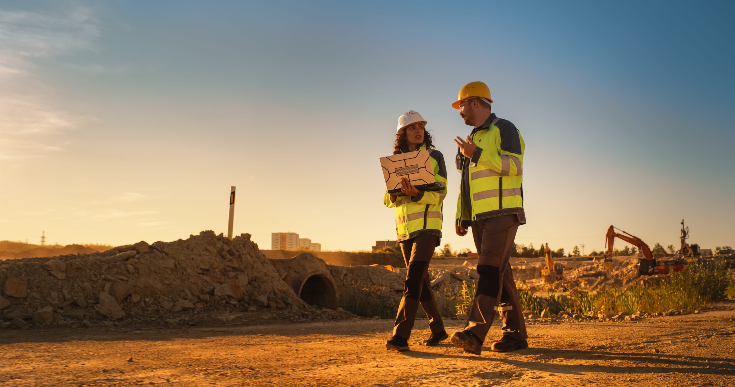 Two construction workers in safety gear discussing plans on a site during sunset.
